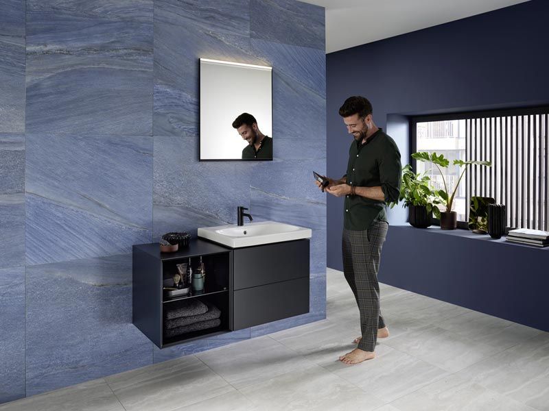 Mann im modernen Badezimmer mit blauen Wandfliesen, dunklem Waschtisch und Pflanzen, lächelt beim Blick auf sein Smartphone.