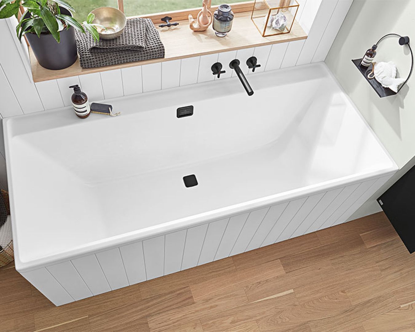 Weiße Badewanne mit schwarzer Armatur vor Fenster und Holzverkleidung.