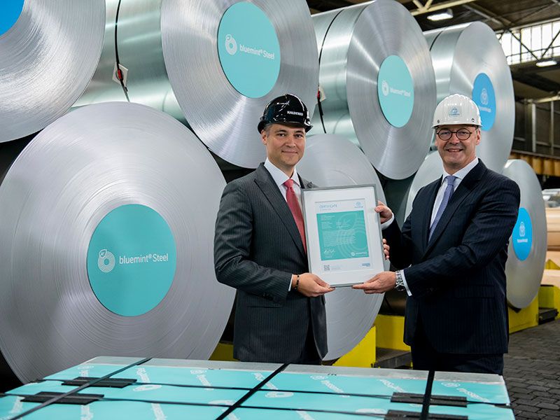 Zwei Männer mit Zertifikat vor Coils aus bluemint® Steel in Produktionshalle.