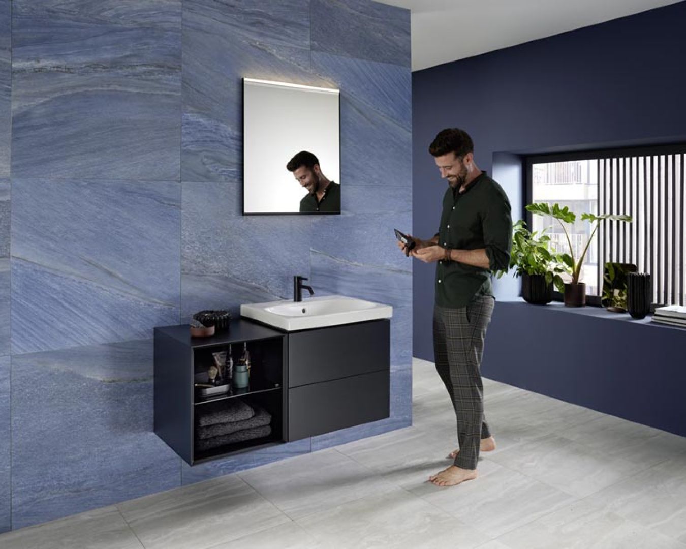 Mann im modernen Badezimmer mit blauen Wandfliesen, dunklem Waschtisch und Pflanzen, lächelt beim Blick auf sein Smartphone.
