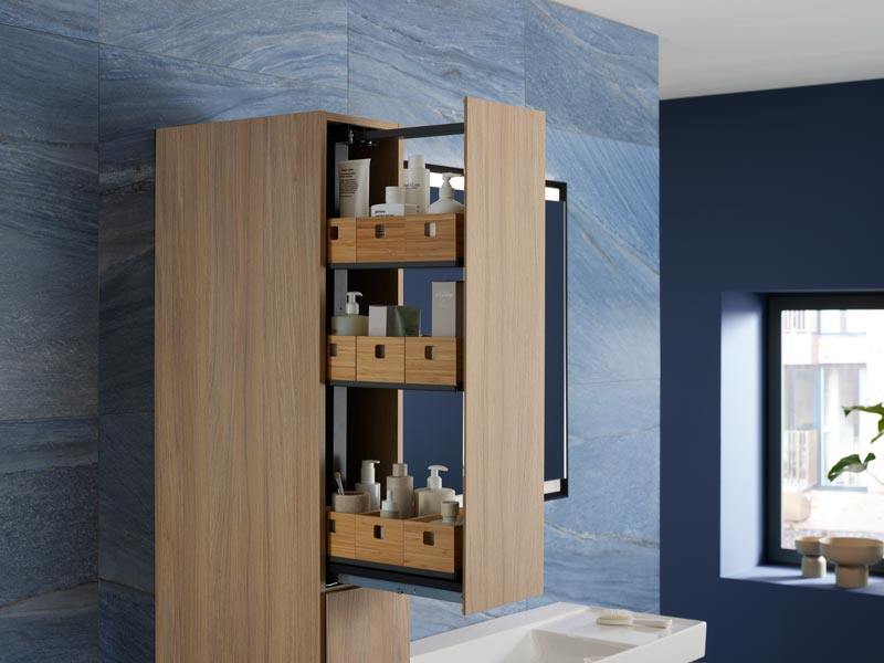 Offener Badhochschrank mit Holz-Auszügen und Pflegeprodukten vor blauer Wand.

